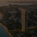 soondri-wongamay-banner2 - Soondri Wongamat Aerial view of a coastal city at sunset featuring a tall modern high-rise near a sandy beach and palm trees along the shore.
