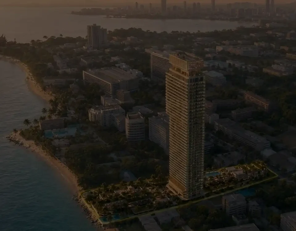 Aerial view of a coastal city at sunset featuring a tall modern high-rise near a sandy beach and palm trees along the shore.